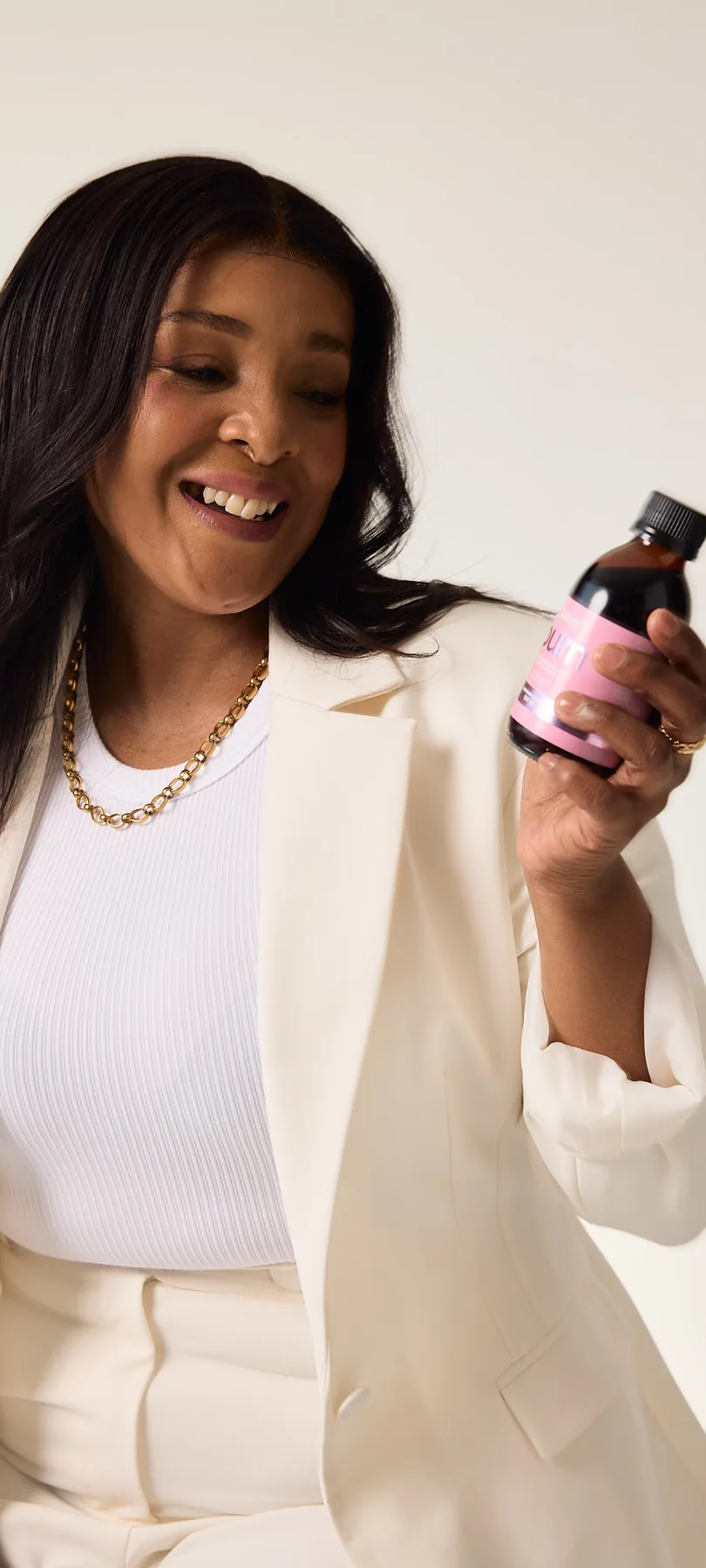 A woman in a cream suit smiles down at a bottle of Sēkret syrup in her hand