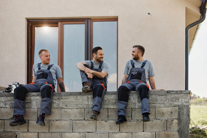 Drei Heizungsbauer sitzen auf der Mauer vor einem Haus und unterhalten sich.