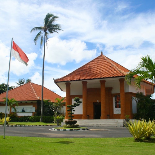 两栋带斜坡屋顶的建筑物,一个挂着红白相间旗帜的旗杆,棕榈树和修剪整齐的绿化,在蓝天下显得格外美丽。