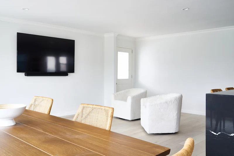 Minimalist dining room with a long wooden table, white chairs, a bench by the window, and a wall-mounted TV. Photo by Todd Huge.