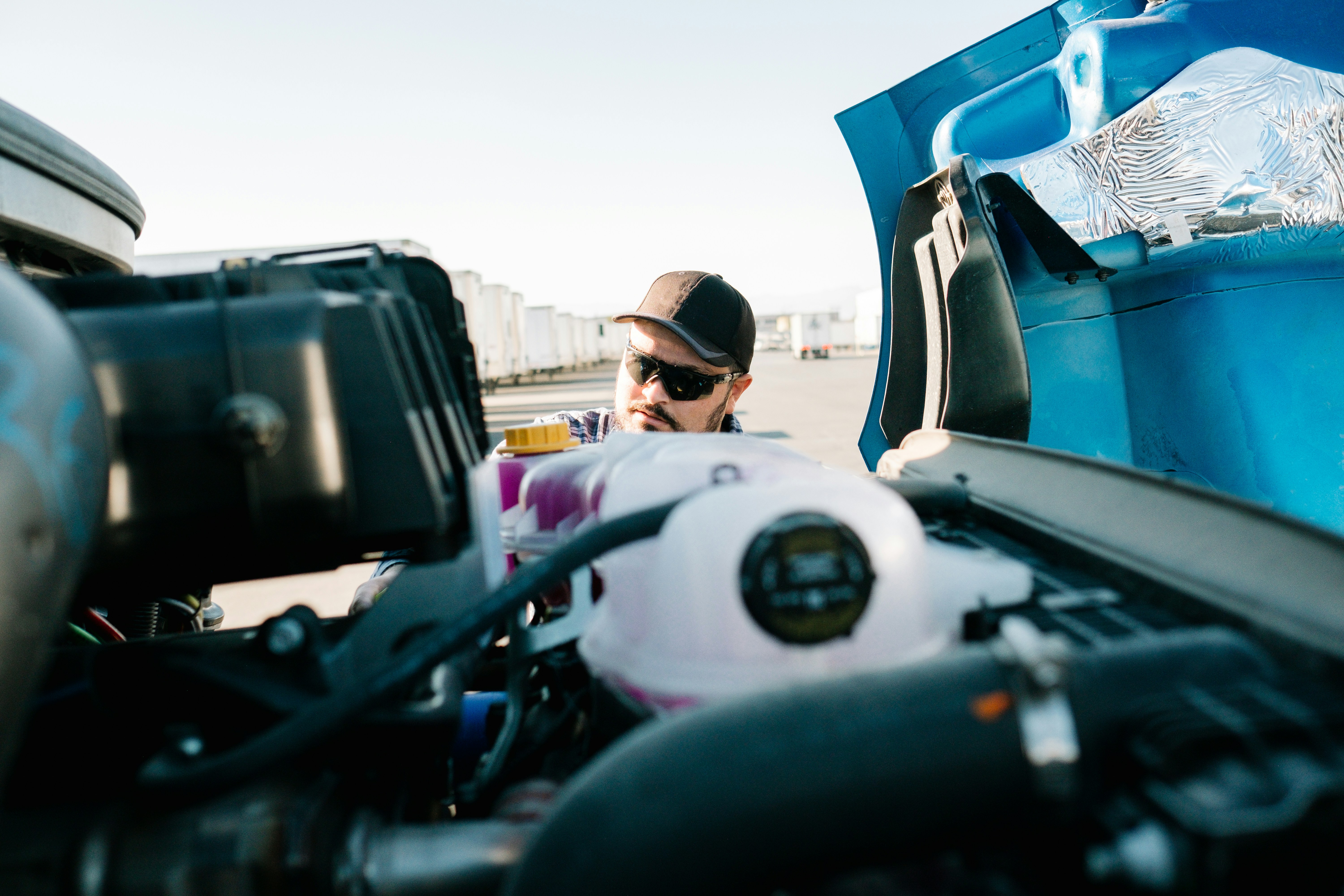 A male truck driver wearing sunglasses and a black cap inspects the engine bay of a blue semi-truck at a commercial trucking yard while he prepares for DOT Week. Shot from a low angle inside the engine compartment, the photo captures fluid reservoirs, wiring, and mechanical components in the foreground, with a row of white trailer units visible in the background. Ideal for content related to truck maintenance, fleet management, pre-trip inspections, and commercial trucking operations.