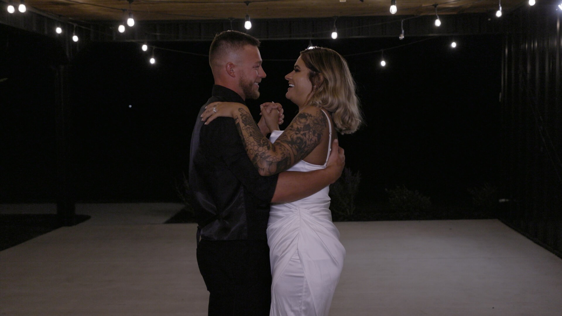 A newlywed couple smiling and dancing on a dimly lit patio on a summer night