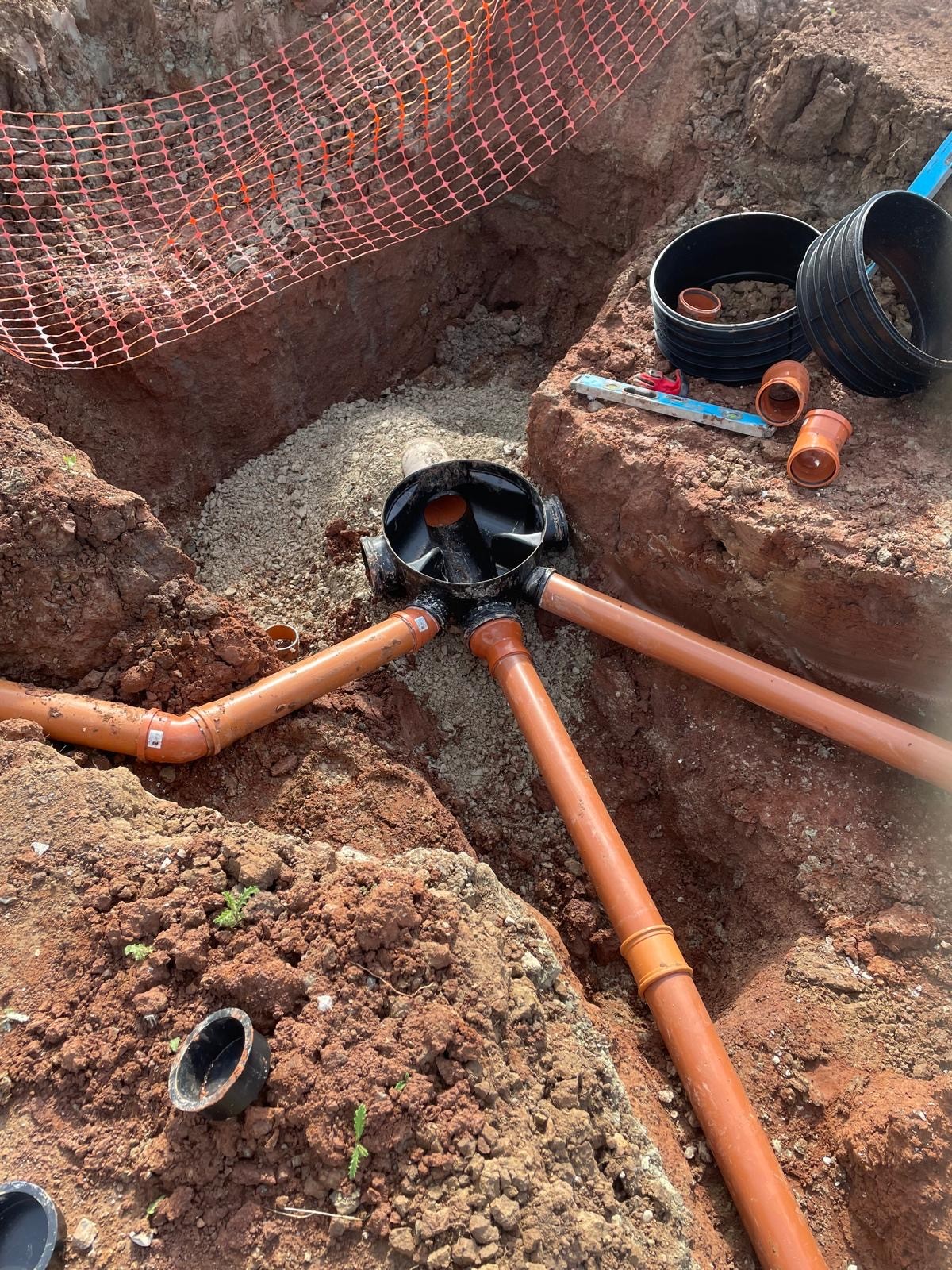 A close-up view of a dug trench showing black pipes connected with orange fittings and tools around the area.