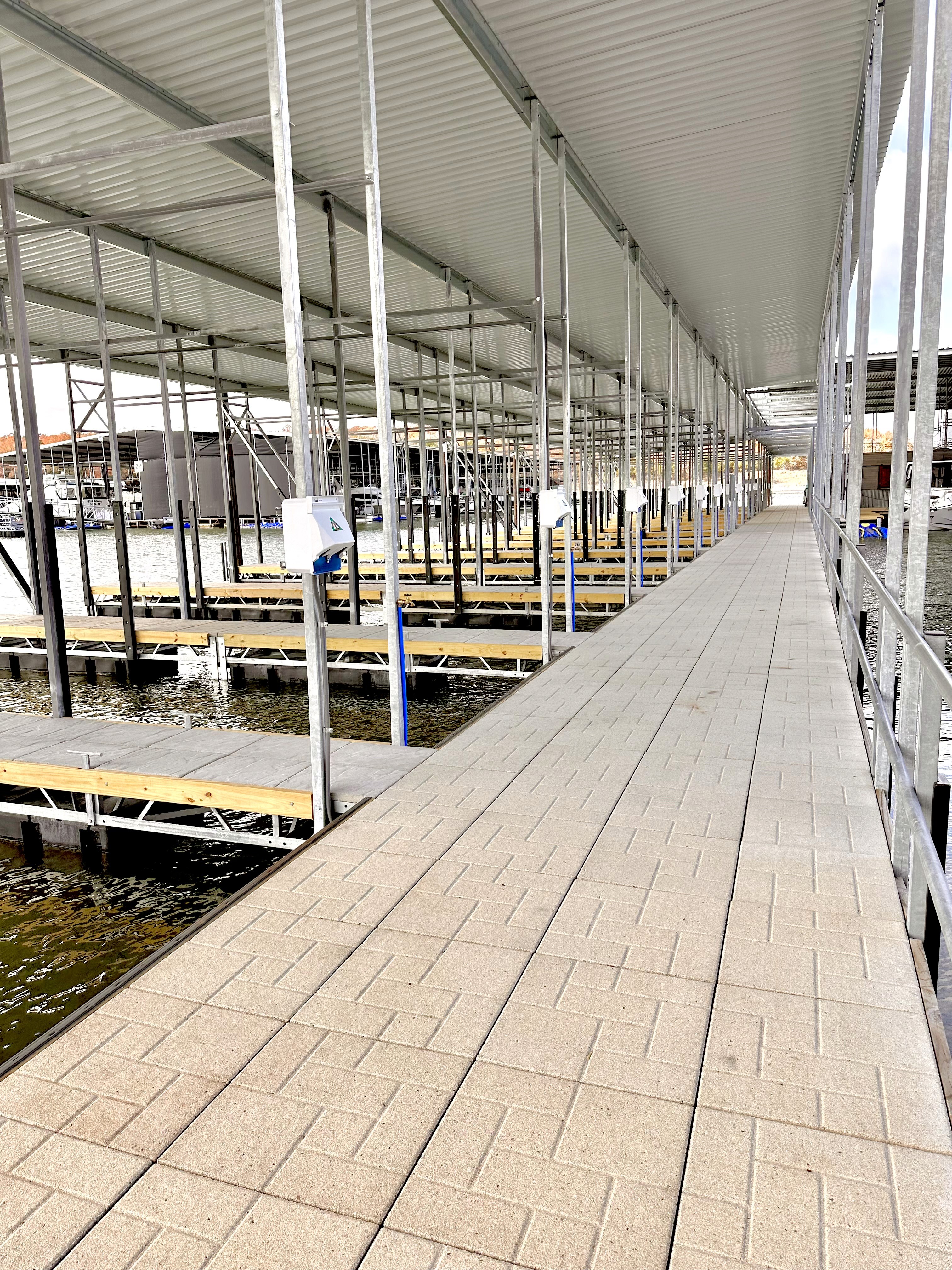 Covered dock area with numerous empty berths and a walkway featuring light brown paving, set against a backdrop of calm water.