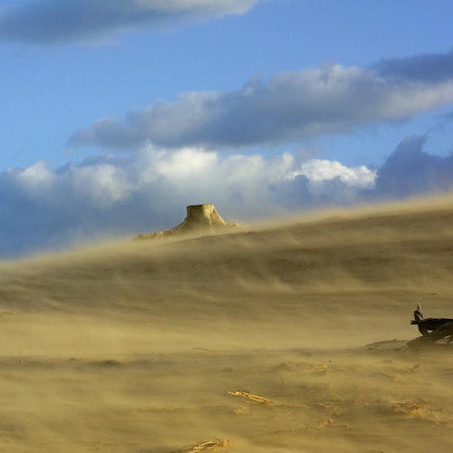 Dusty desert landscape with a butte in the distance under a blue sky with clouds.