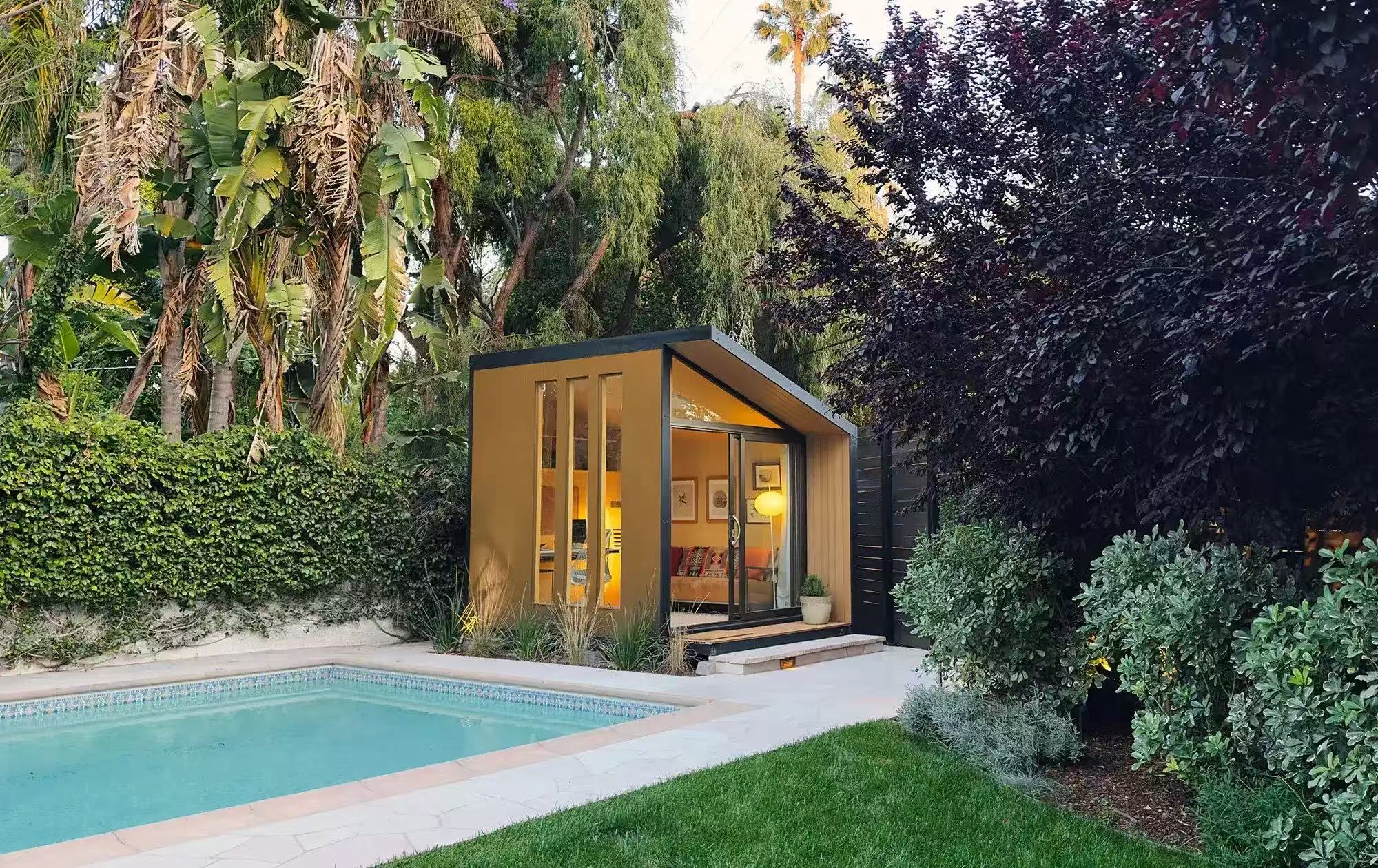 Modern luxury house on a hillside surrounded by trees, featuring large glass windows, a terrace, and a small pool at sunset.