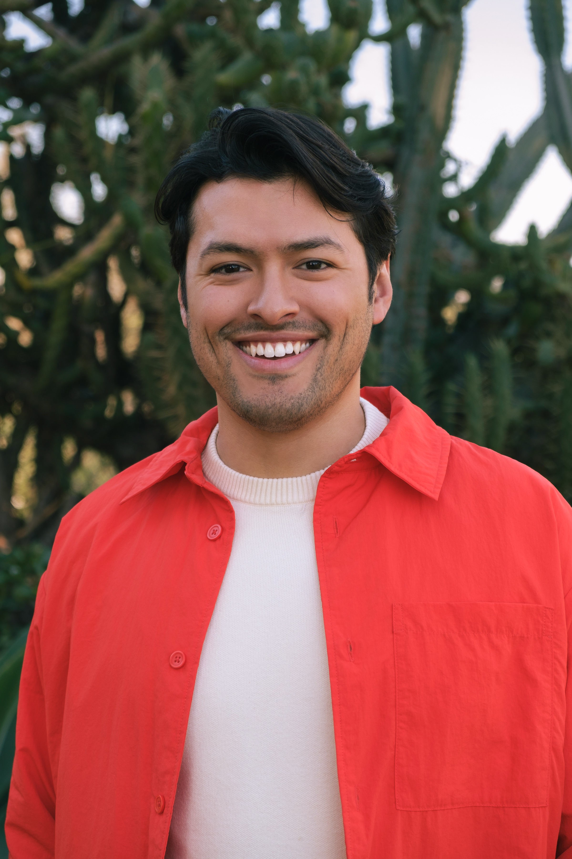 A young man with dark wavy hair and facial stubble smiles broadly at the camera. He appears to be of Latino heritage and is wearing a bright coral-red button-up shirt over a cream-colored ribbed sweater or top. The background shows lush green foliage with dappled sunlight filtering through the leaves, creating a natural outdoor setting. His expression is warm and genuinely happy, and the lighting creates a soft, golden hour effect on his face.