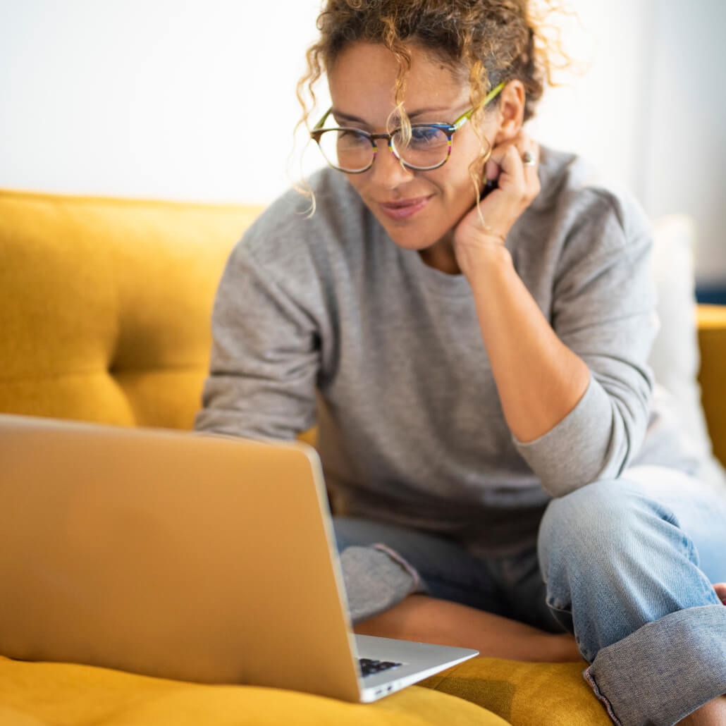 Woman on a yellow couch works on a laptop.