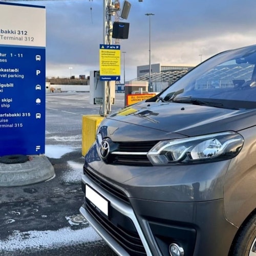 Una furgoneta gris está estacionada en un aparcamiento cerca de señales azules y amarillas que indican direcciones para aparcamiento y terminales. El cielo nublado se ve al fondo.