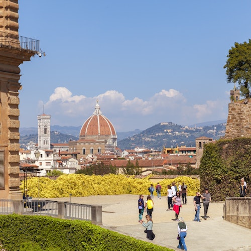 People walking in a garden with Florence's Duomo and bell tower in the background, under a clear blue sky.