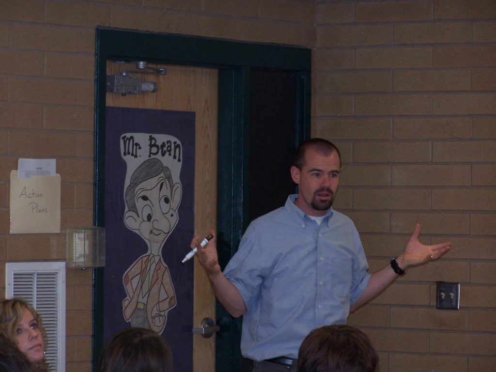 A man stands in front of a classroom gesturing with a marker in hand. Behind him, a cartoon poster of Mr. Bean is taped to a wooden door. A paper labeled “Action Plans” is on the wall.