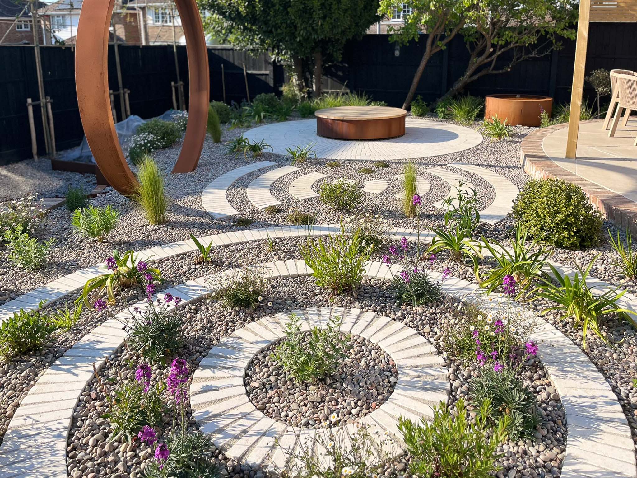 A landscaped garden features circular stone pathways surrounded by greenery and a central area for seating.
