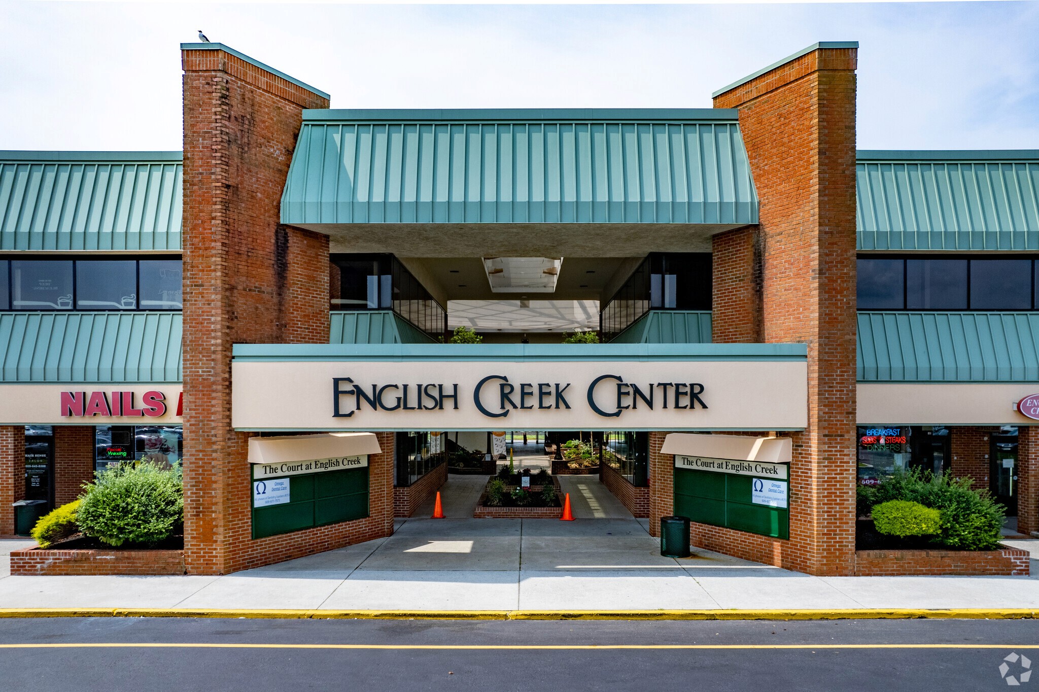 Brown brick shopping center with a green metal roof, featuring a sign reading "English Creek Center." Entrances to various shops are visible.