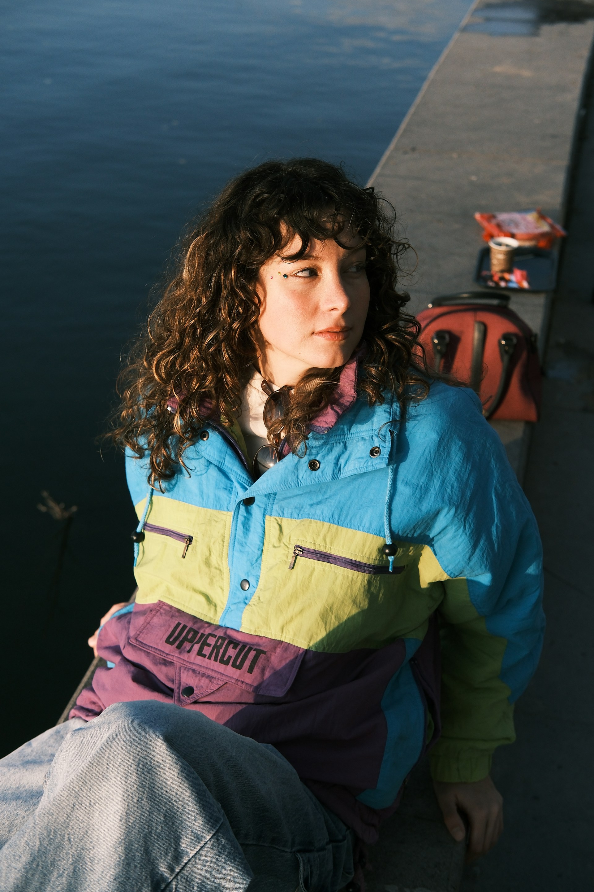 A woman in a blue, lime green, and purple "Uppercut" windbreaker sitting on a stone ledge by the water. She is looking off to the side with a calm expression.