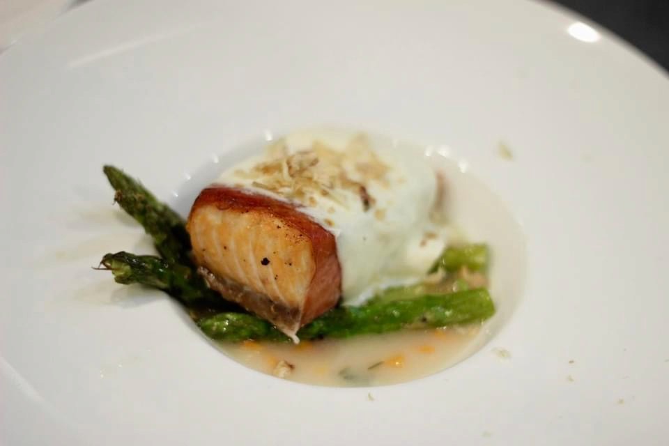 A white plate displays a salmon fillet on asparagus,  topped with white sauce and garnish