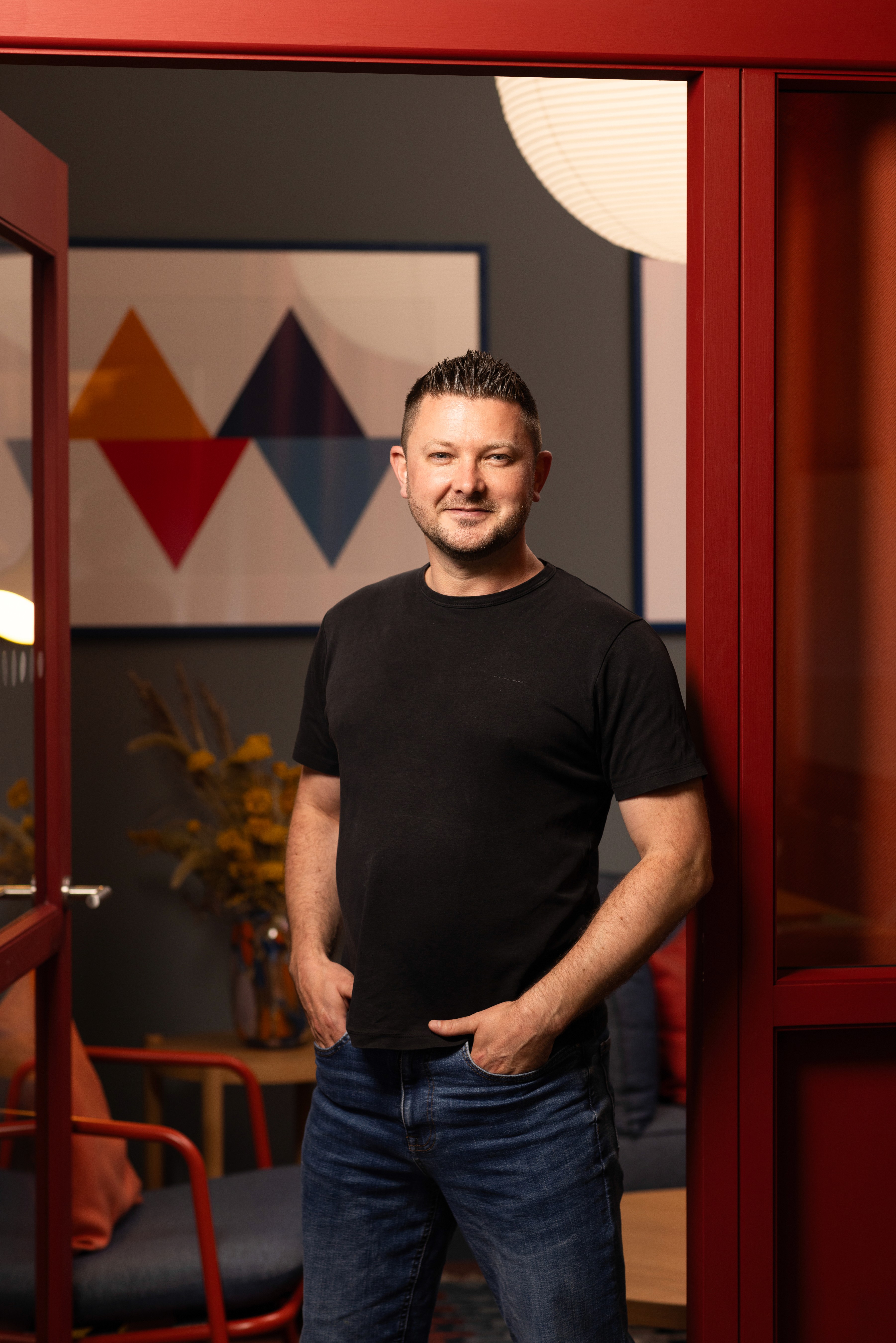 Photo of David Sargison smailing at the camera in an open doorway wearing  black t-shirt and blue jeans with his hands in his pockets
