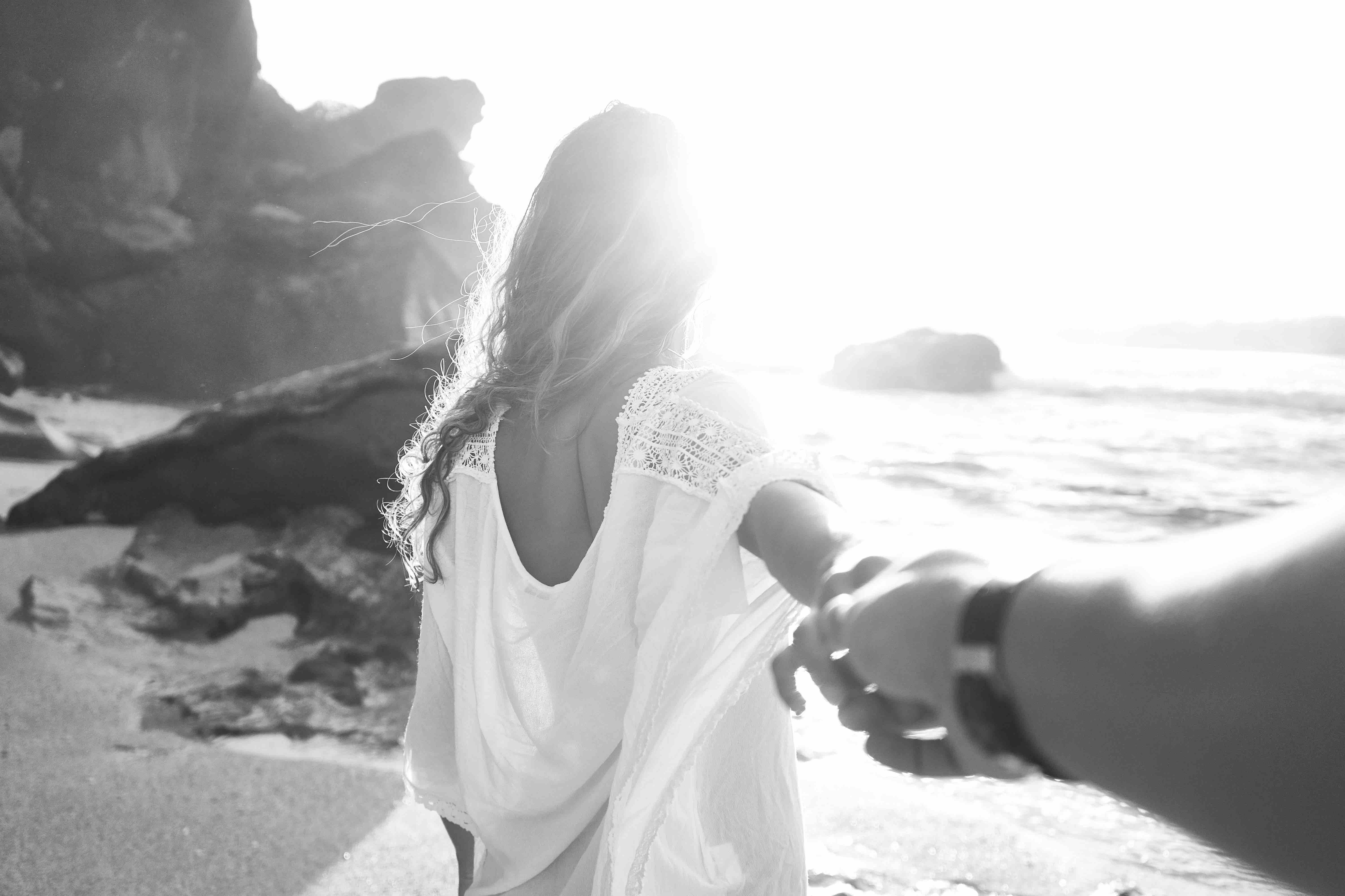 Perspective shot of male and female couple at the beach, focusing on the female in a flowing beach dress while she holds his hand, leading him forward into the blurred out ocean backdrop