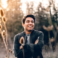 A smiling young man with dark hair stands outdoors with his arms crossed, wearing a dark jacket over a gray shirt, surrounded by tall dried plants and trees with soft sunlight filtering through.