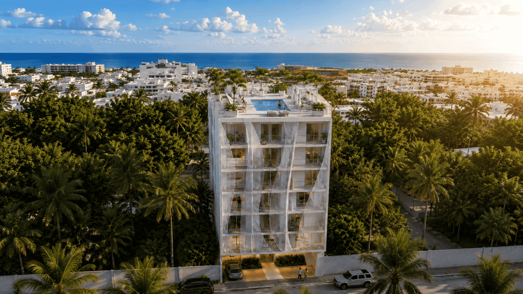 Vista aérea de edificio residencial blanco con fachada ondulada, rooftop con alberca y el mar al horizonte entre vegetación tropical