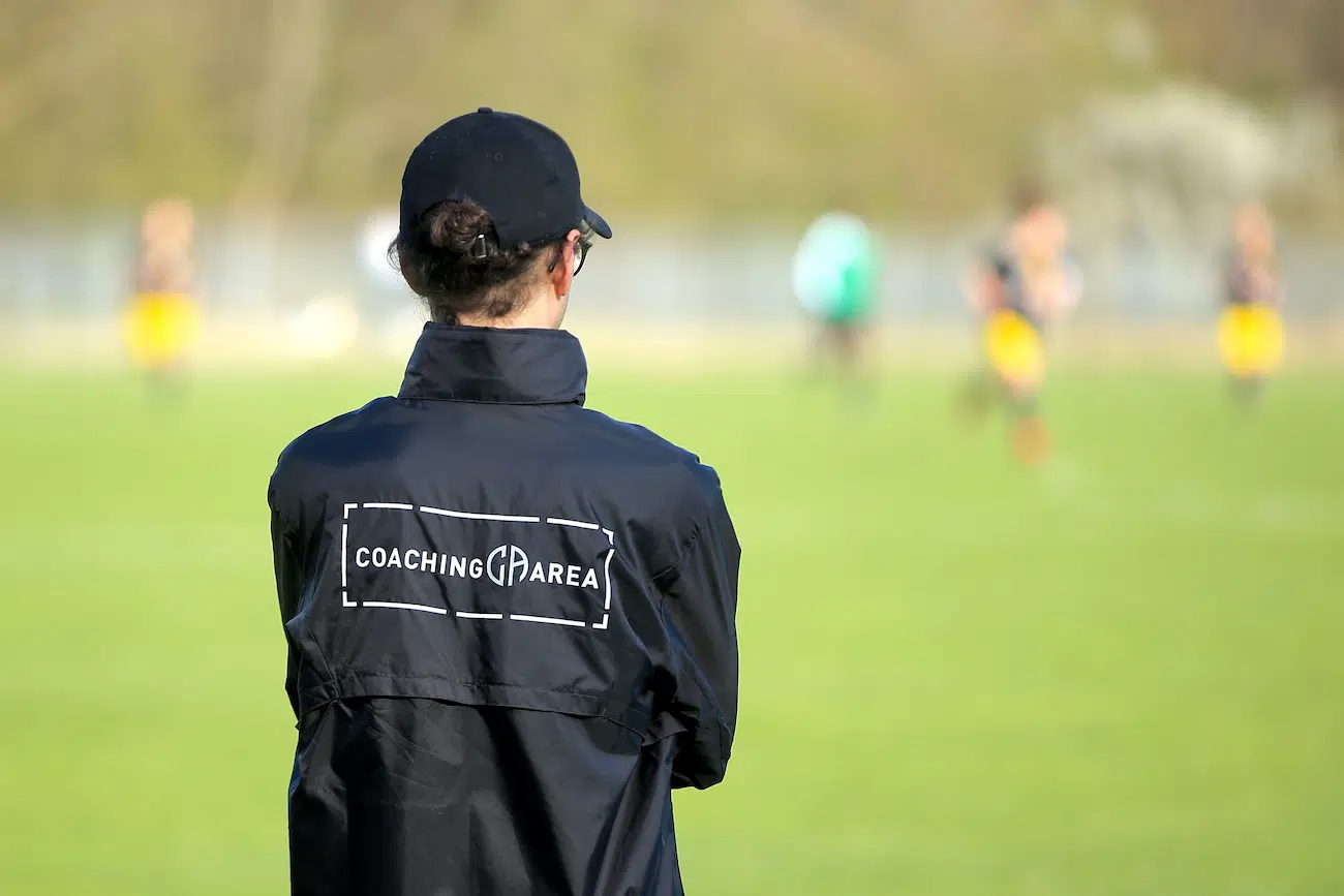 A football coach wearing a CoachingArea-branded jacket and cap observes a match on a grassy field. In the background, players in action are visible but blurred, emphasizing the coach's role in team management and strategy. The image represents the essence of connecting with other teams and finding the right opponents for matches, tournaments, or leagues through CoachingArea.