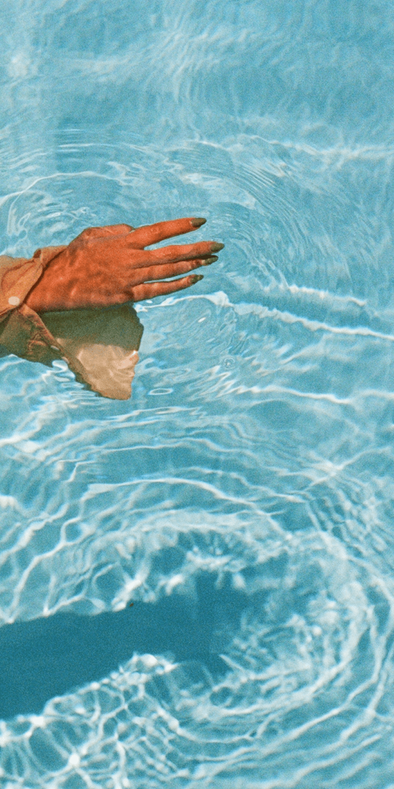 A person wearing a sheer, long-sleeved garment extends their arm into a clear blue pool, creating gentle ripples in the water with sunlight reflecting on the surface.