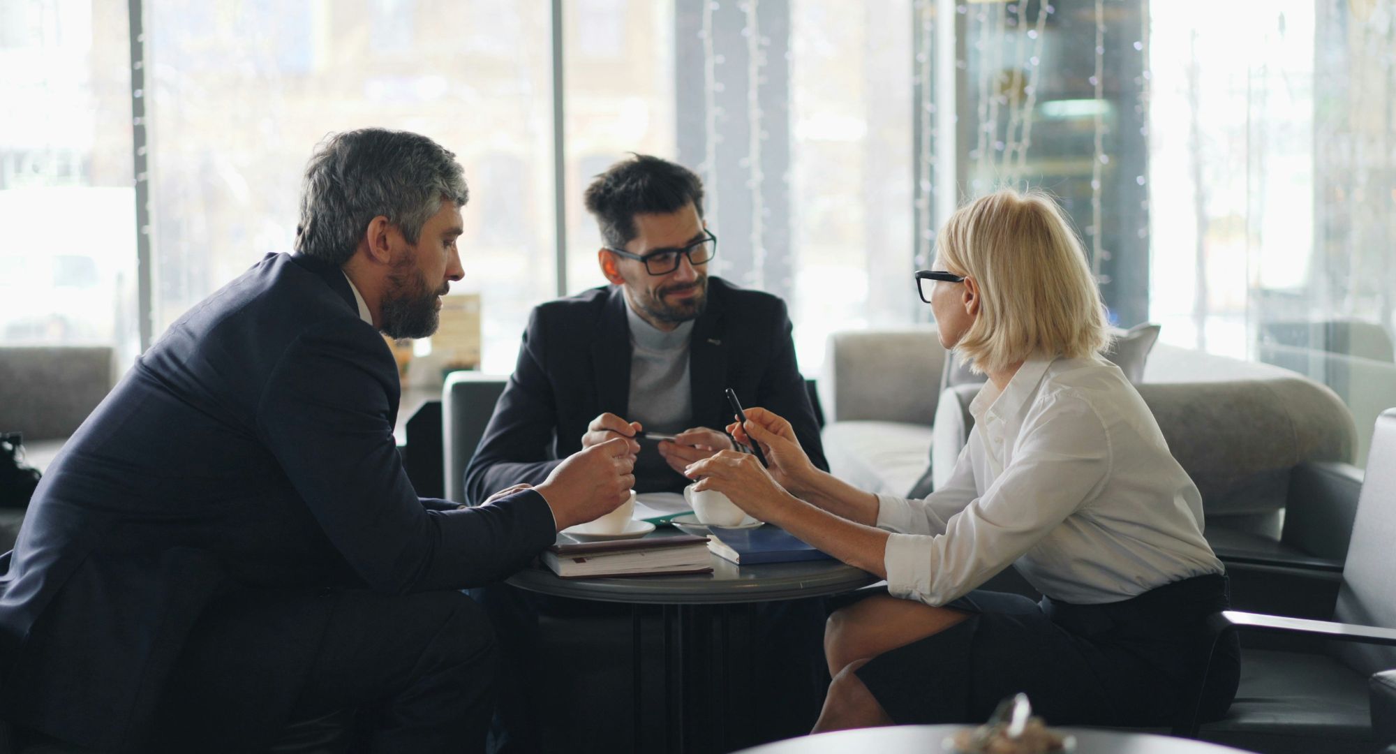 dois homens e uma mulher conversando sobre trabalho em um escritório num andar alto do prédio com janelas de vidro mostrando outros prédios ao fundo