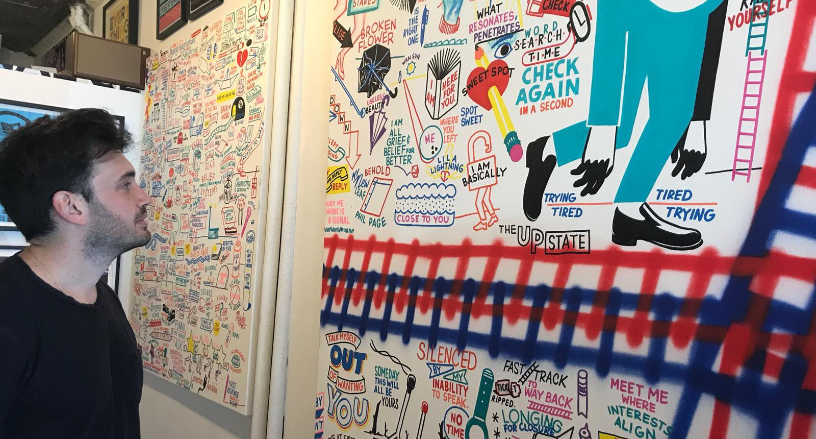 A photo of a man looking at a white wall filled with colorful doodles.