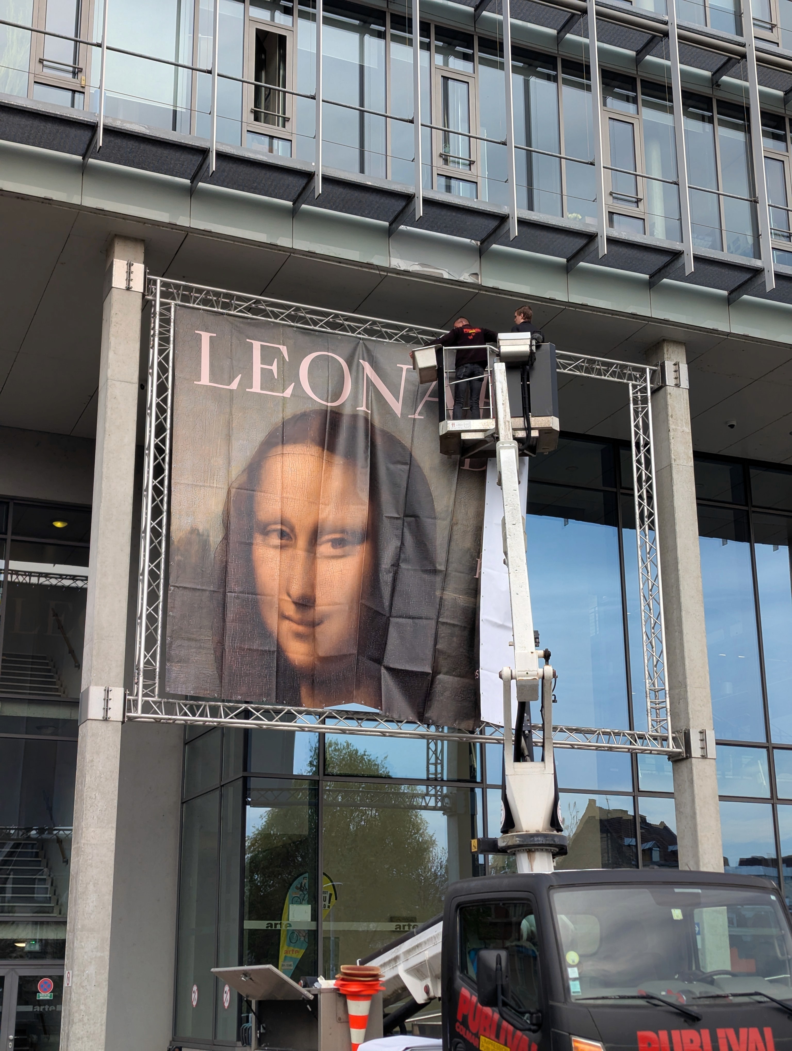 A large banner featuring a portrait of Mona Lisa is being hung on a modern building by workers in an aerial lift.