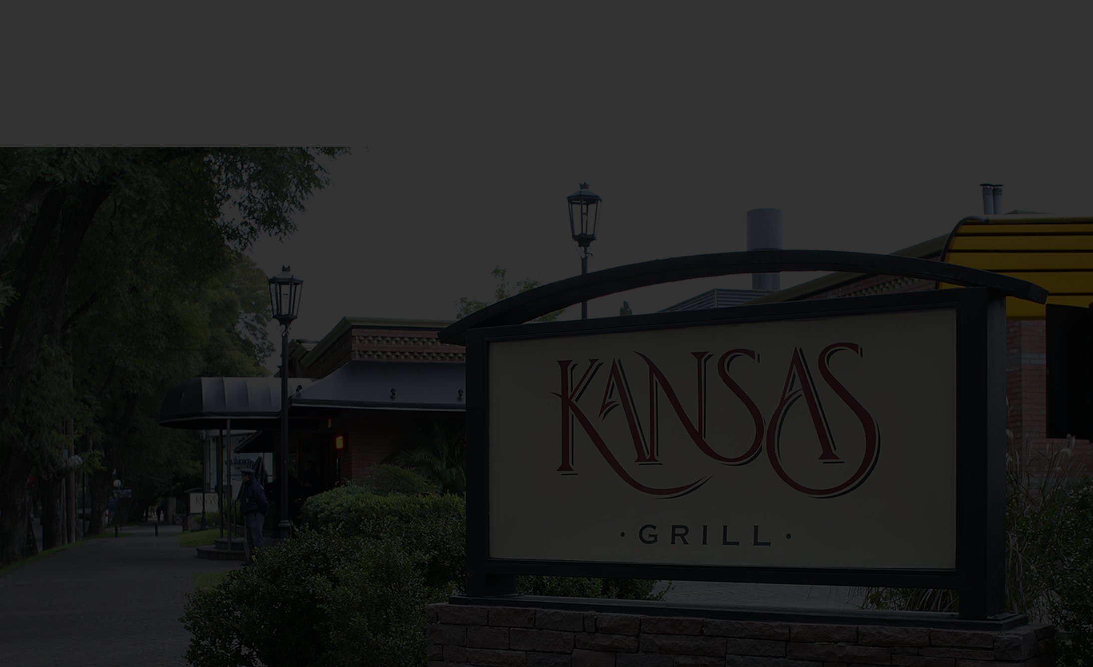Sign for "Kansas Grill" with elegant lettering, set in a lush, landscaped area with trees and lampposts. The scene is calm and inviting.