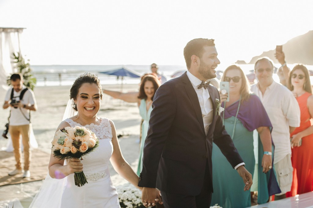Beaming bride holding a peach and succulent bouquet as she walks with the groom past cheering guests at a beach wedding