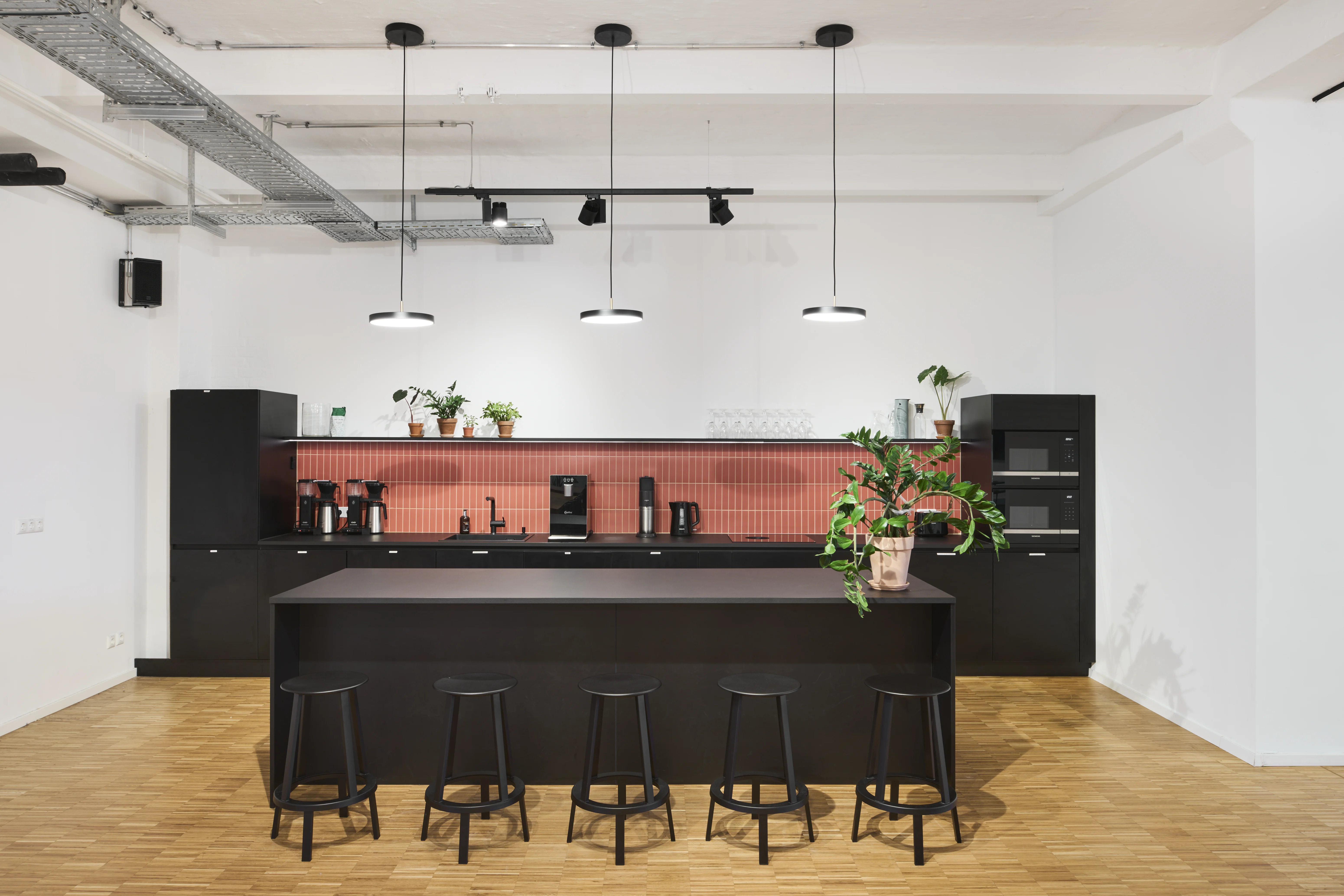 Black kitchen at PLAN A with a long black island, black bar stools, coral-red tiled splashback, plants on the counter and three black pendant lights.