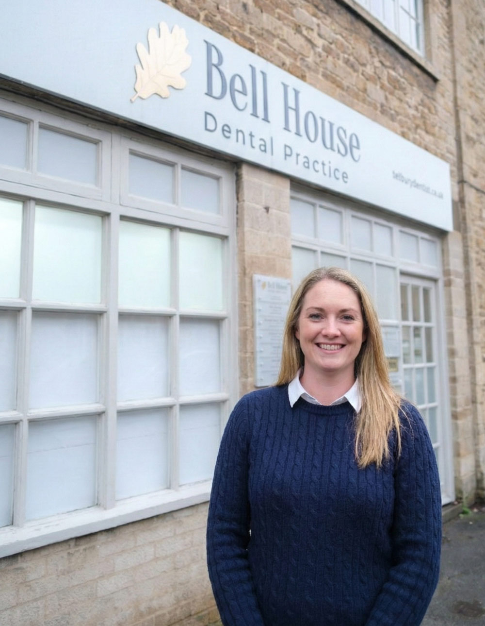 A smiling individual stands in front of Bell House Dental Practice, a building with large windows and a brick exterior.
