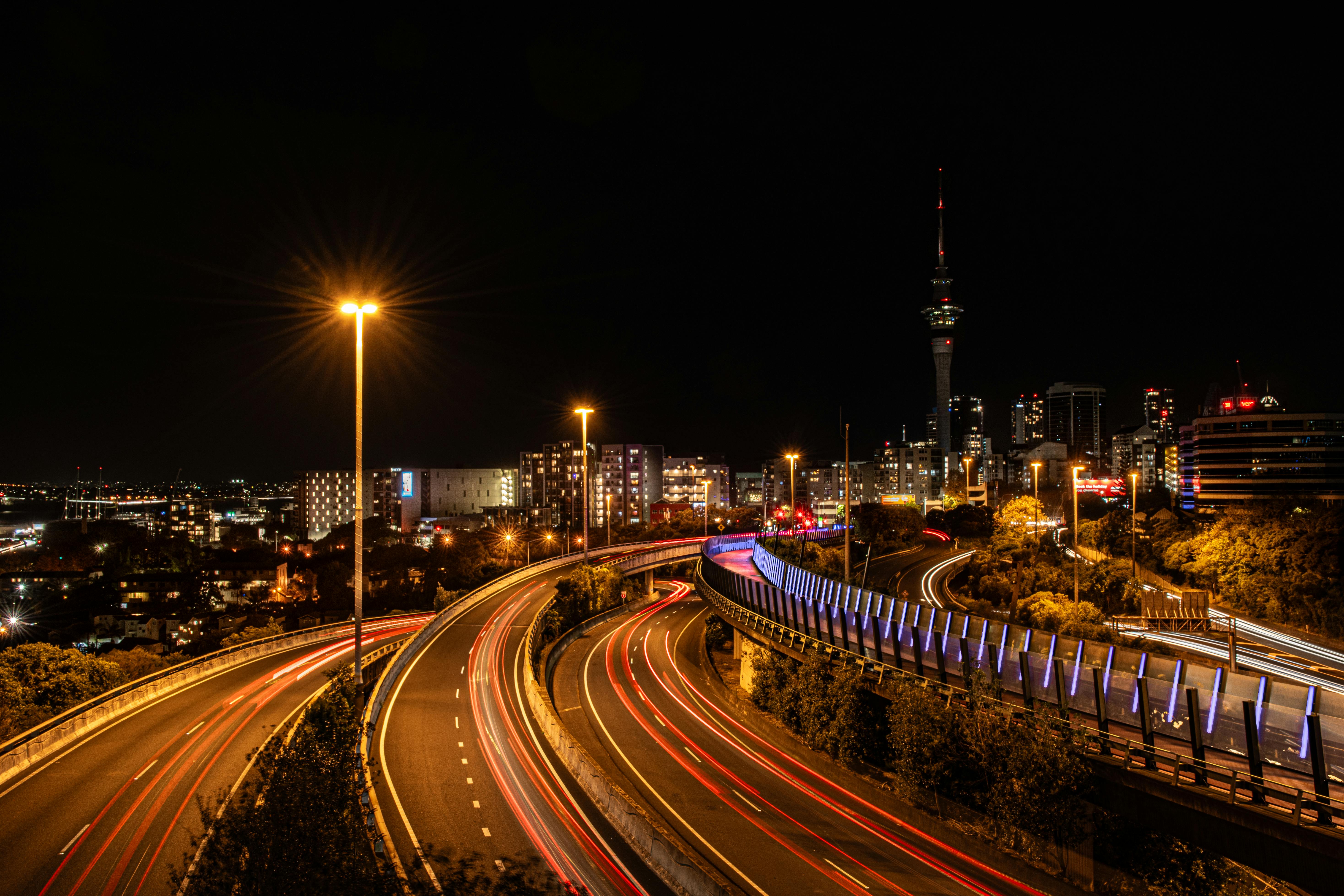 A long-exposure night shot of a multi-lane highway in Auckland. Streaks of red and white light from moving cars create a sense of motion, leading the eye toward the illuminated Sky Tower and city skyline in the distance.