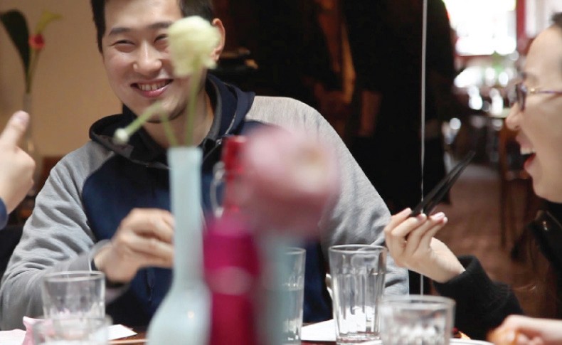 Two people are seated at a table. The man on the left wears a gray hooded sweatshirt and smiles at the camera. The woman on the right wears glasses and holds a pair of dark chopsticks. Several clear drinking glasses are arranged on the table in front of them. Behind them, a blurred figure is visible. The table is decorated with several vases containing pink and white flowers.