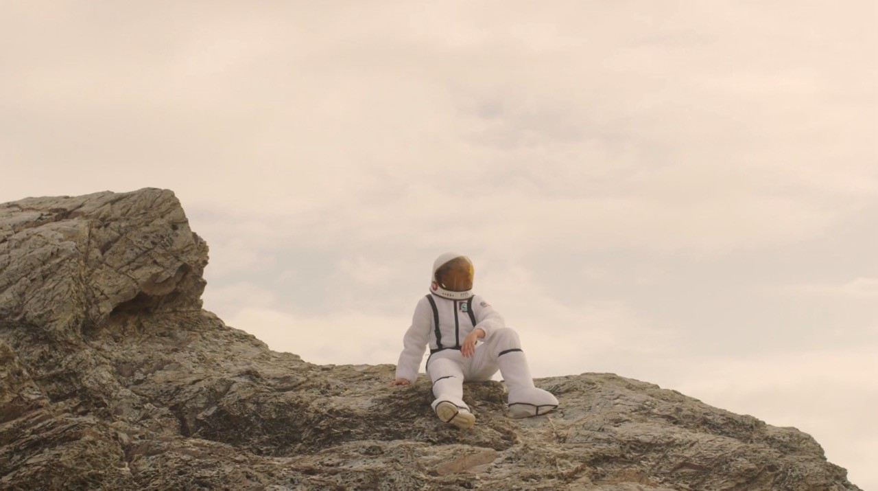 Person in a white spacesuit sitting alone on a rocky outcrop under a soft, cloudy sky.