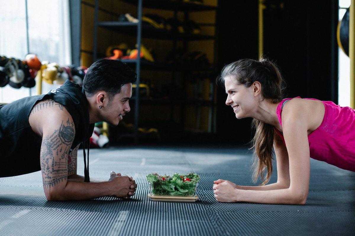Two people doing planks face off with a bowl of salad between them.