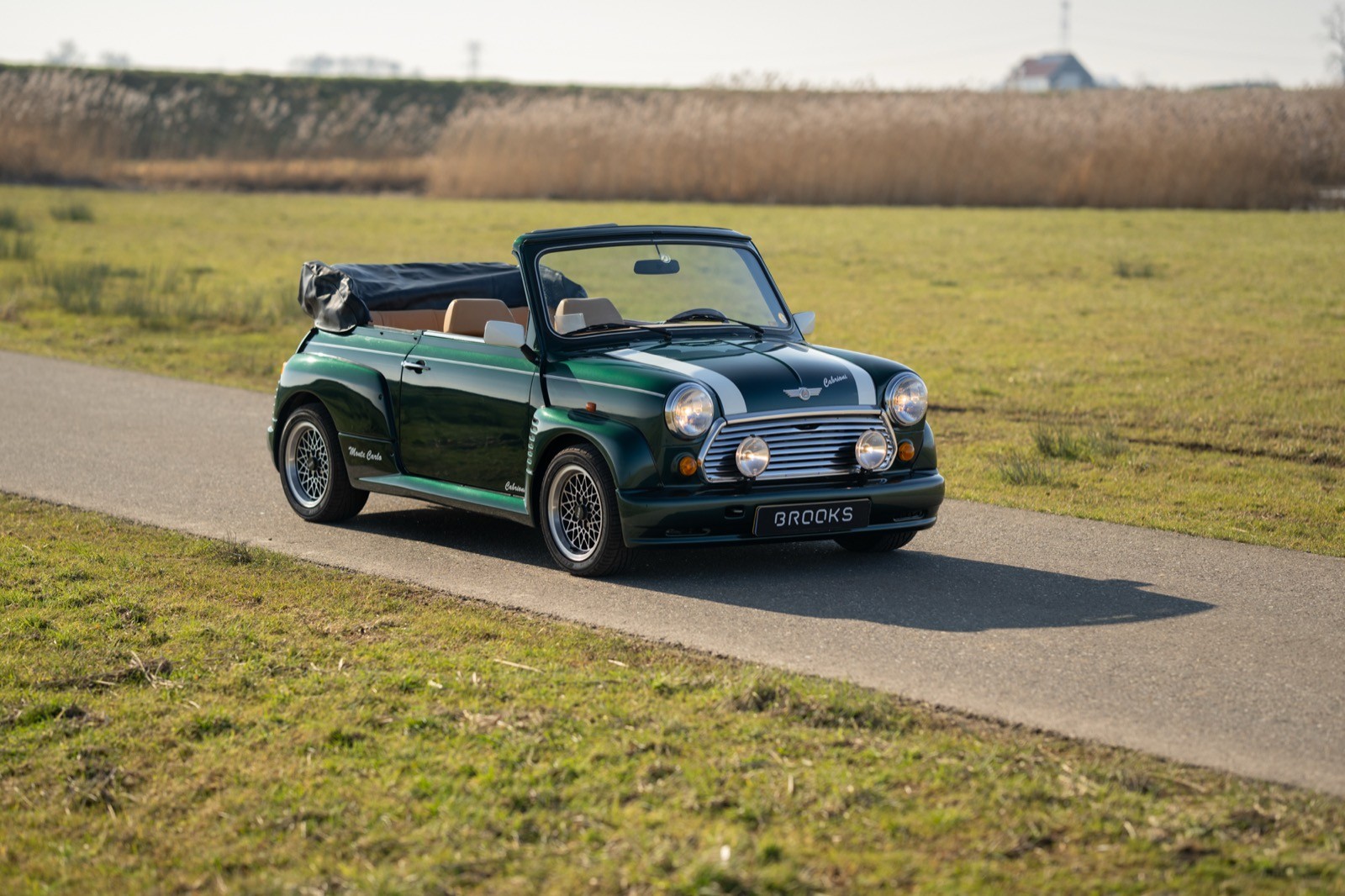 A green convertible car driving along a country road, surrounded by grassy fields under a clear sky.