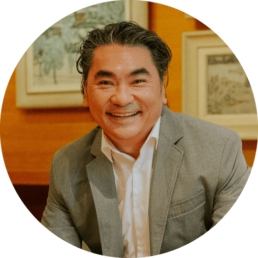 A person in a gray suit and white shirt smiles at the camera against a warm-toned background with framed artwork.