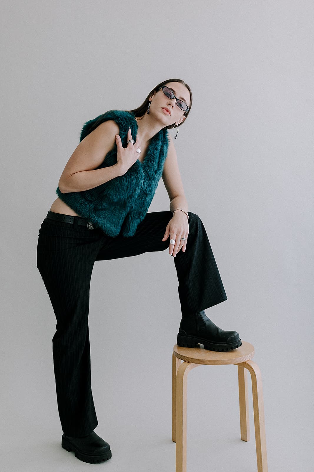 A model poses confidently with one foot on a stool, wearing a teal fur vest and black pants, in Revelator Studio, a natural light photography studio in Shreveport.