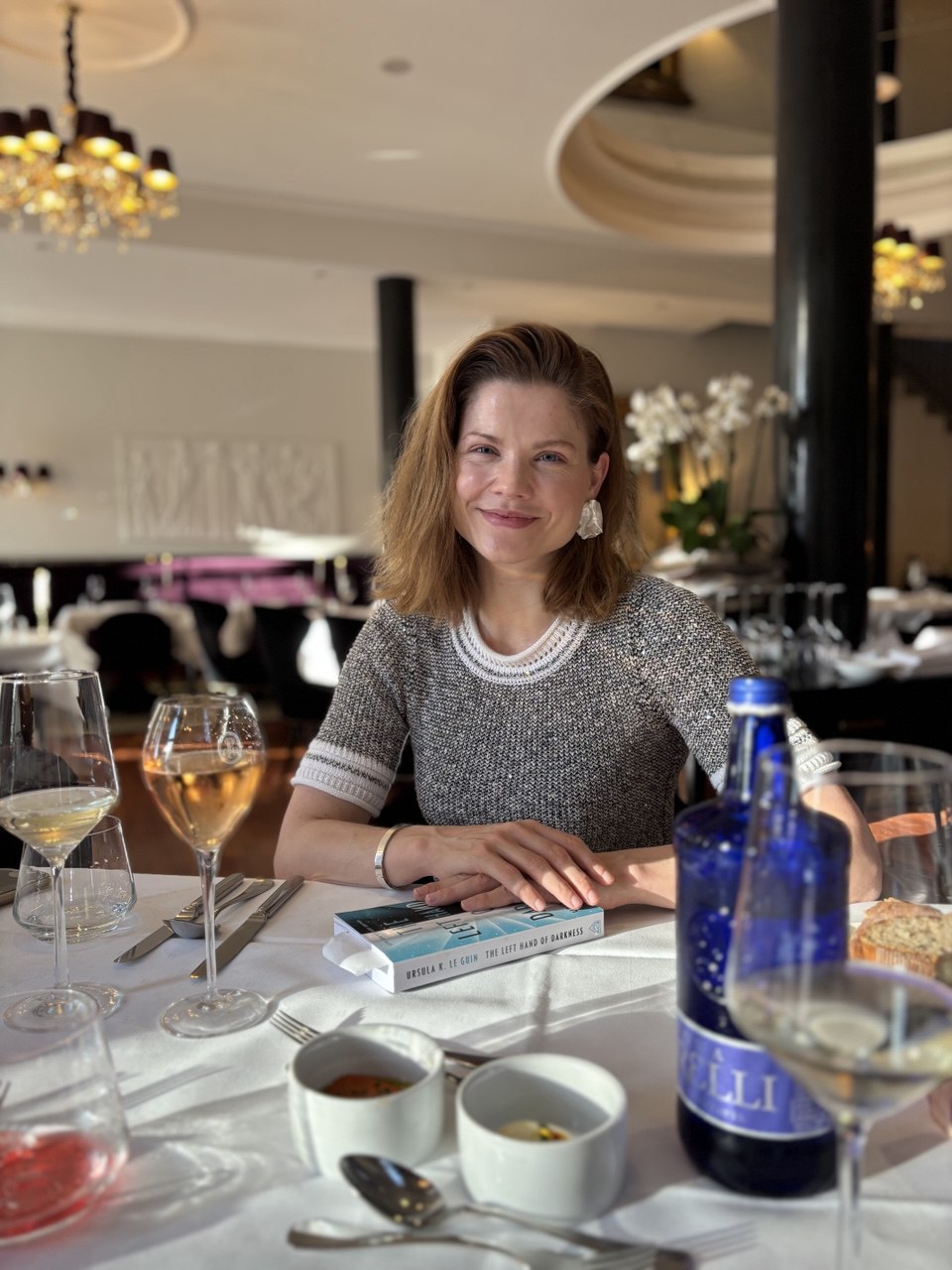 Tosca de Jong at a restaurant table with wine glasses, smiling in soft light, with Ursula K. Le Guin’s book *The Left Hand of Darkness* in front of her.