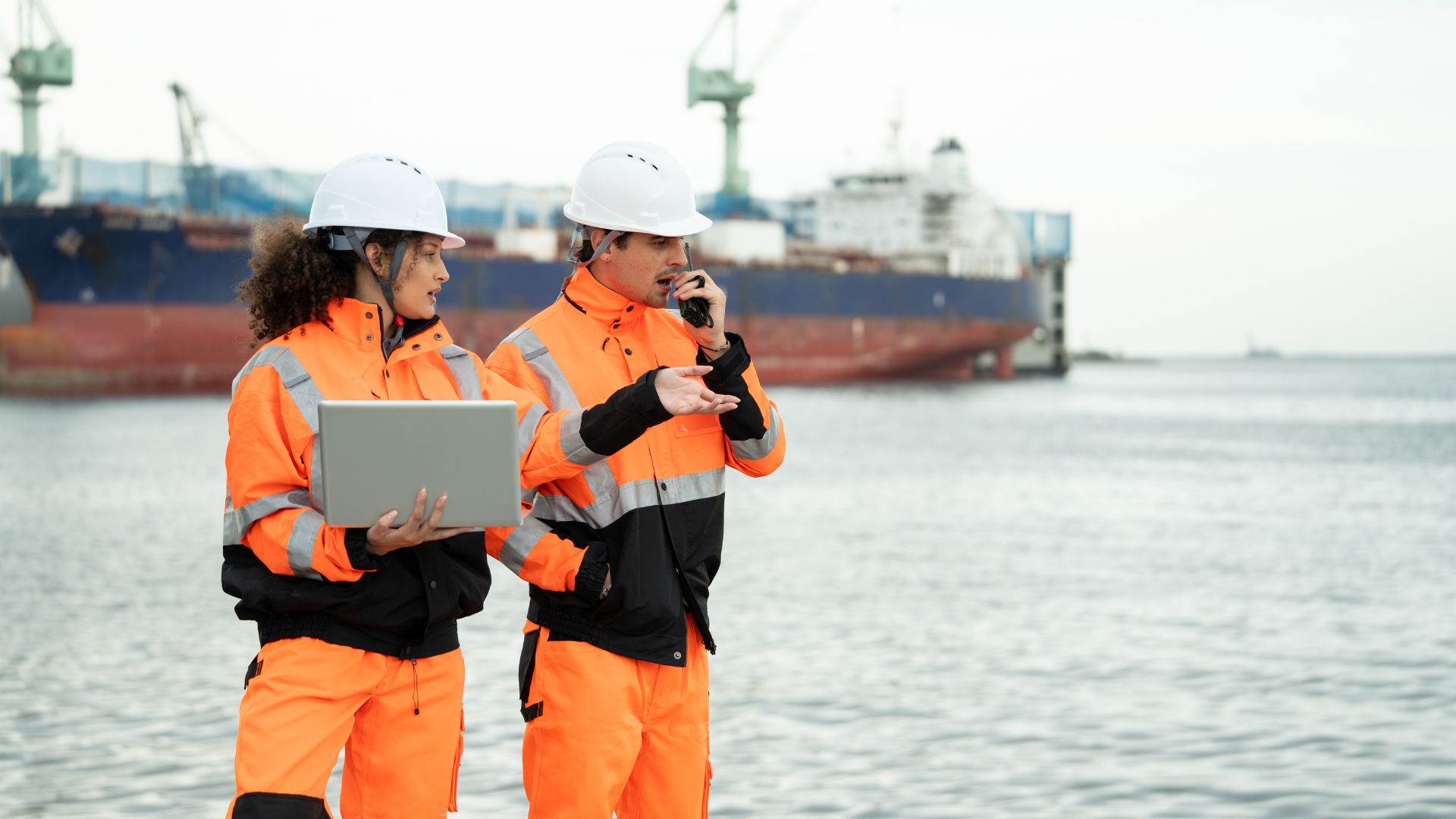 Two individuals in orange workwear and hard hats are standing by the water. One holds a laptop, and the other speaks into a walkie-talkie, with a large ship docked in the background.