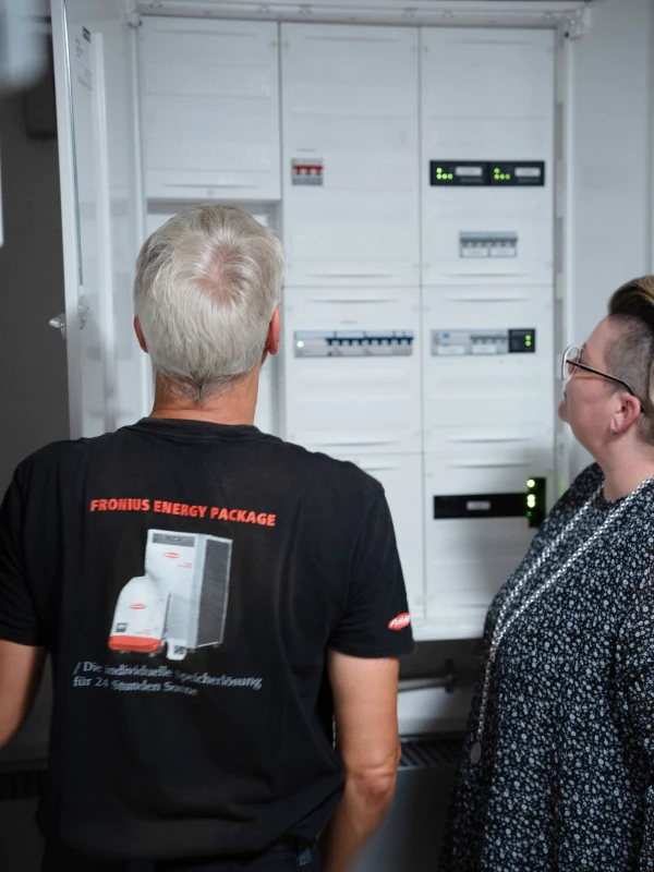 Handwerker Fersing und Eigentümerin Riedesel-Nüßgen im Beratungsgespräch im Keller vor dem Zählerschrank und dem PV-Speicher