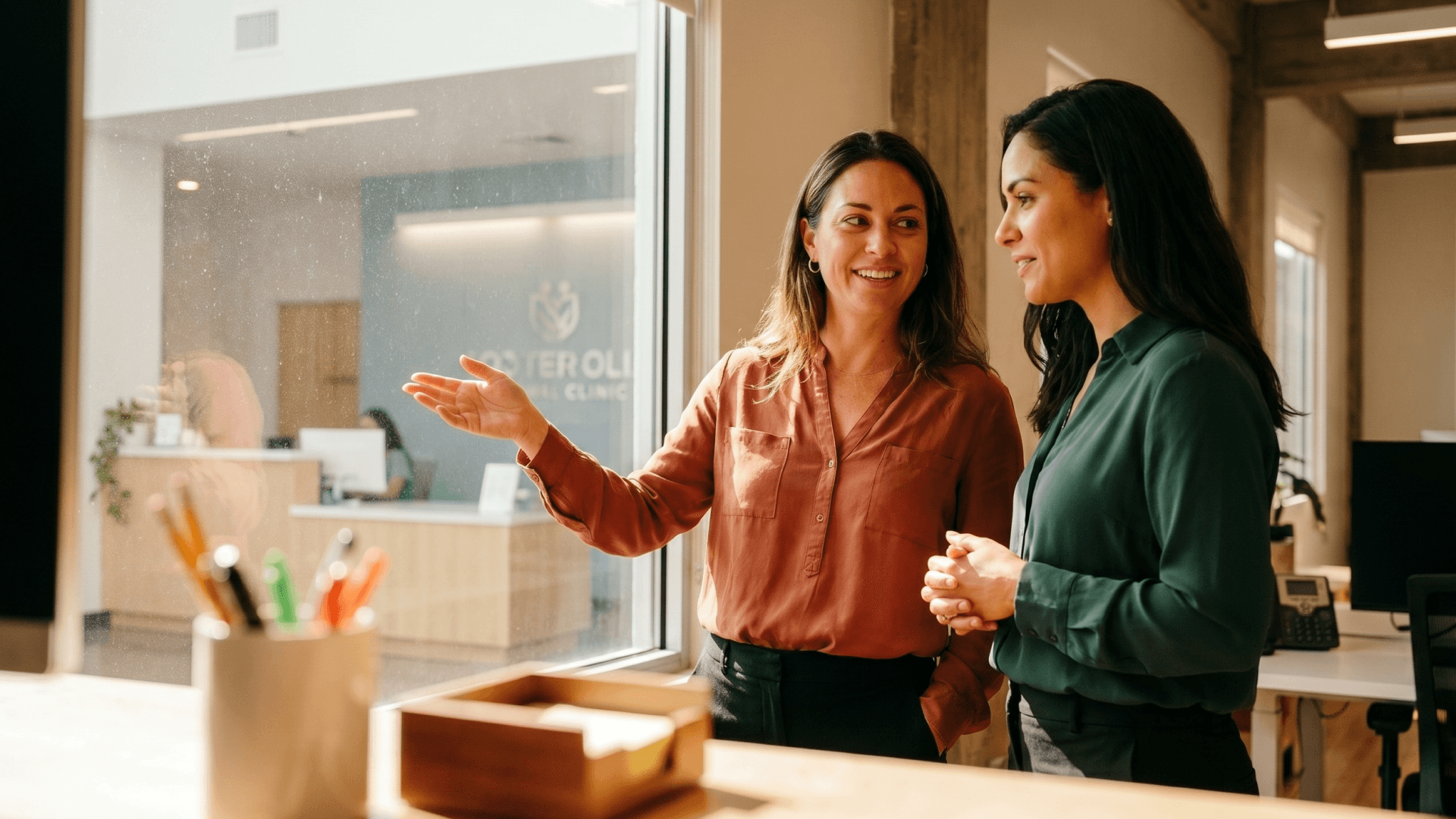 Duas mulheres conversam de pé em um ambiente de recepção iluminado por luz natural, uma de blusa terracota gesticulando enquanto explica algo à outra, de camisa verde, ao fundo o letreiro de uma clínica em parede de vidro, representando o momento de onboarding e acolhimento que a Tivita apoia nas clínicas.