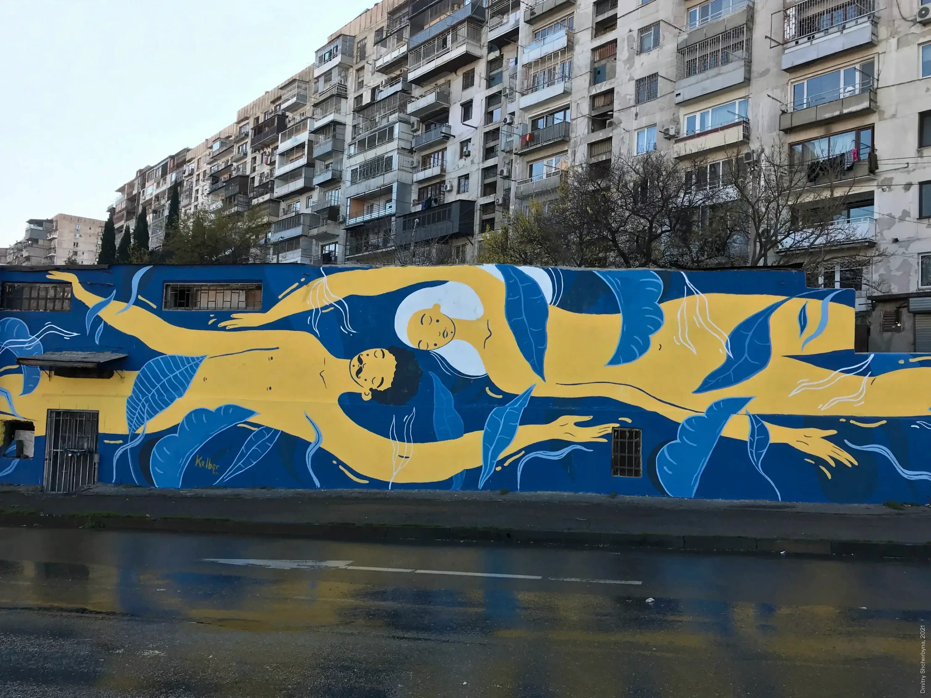 A long horizontal mural of stylized human figures floating and intertwining in blue and golden yellow, painted on a low wall in front of a Soviet-era apartment building.