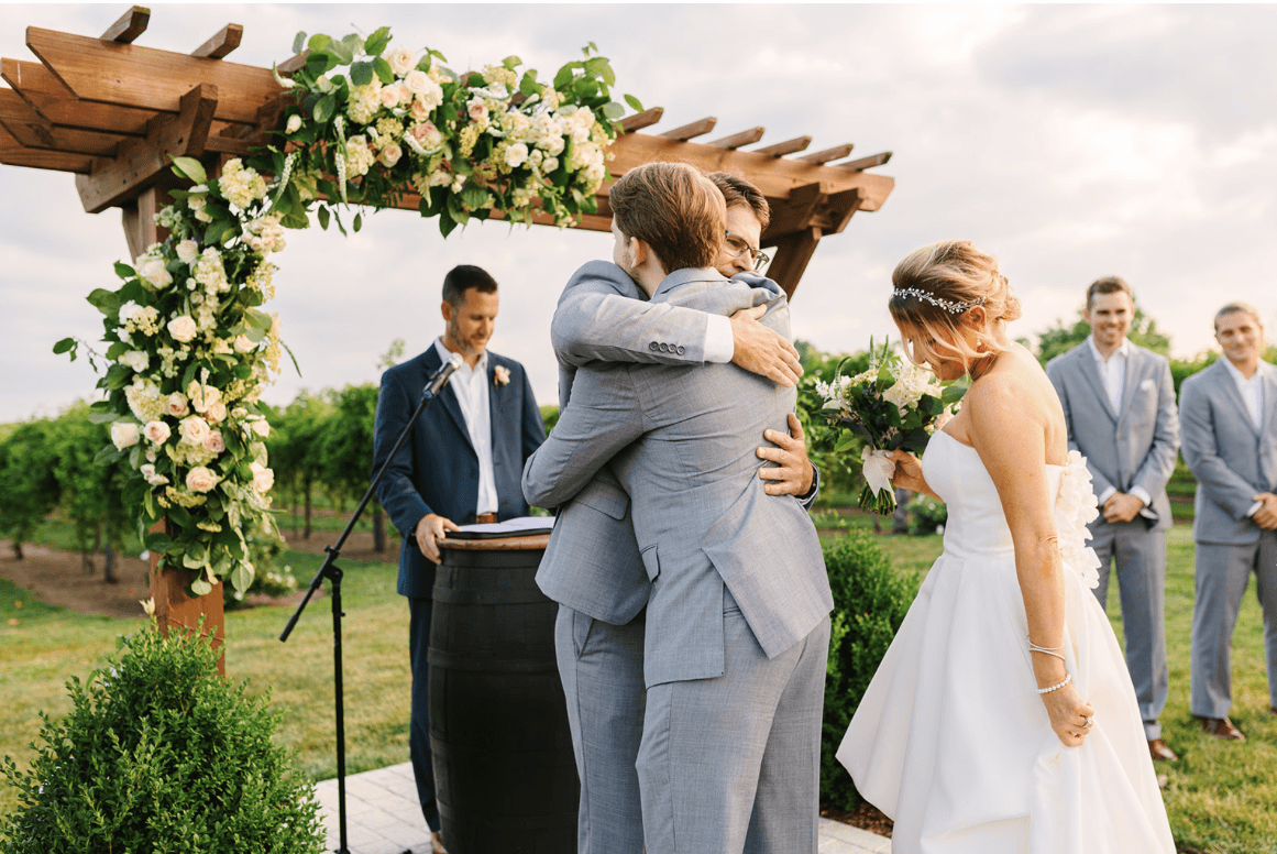 Outdoor wedding ceremony under a wooden pergola decorated with greenery and white flowers. The groom in a gray suit hugs another man as the bride in a strapless gown smiles, holding her bouquet. Groomsmen in gray suits stand in the background, and the officiant watches nearby.