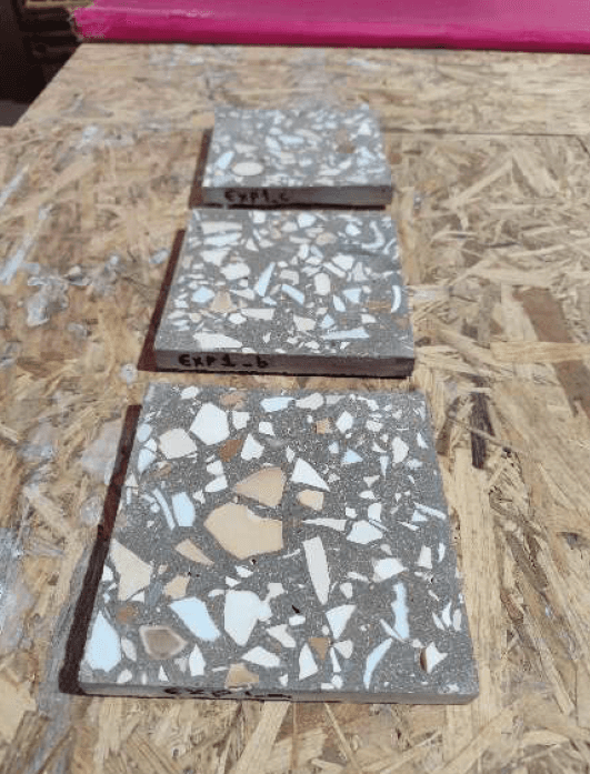 Trois carreaux de terrazzo expérimental étiquetés EXP1-C, EXP1-B et EXP1-A, posés sur une planche en OSB, présentant différentes densités d'éclats de céramique blanche et beige sur fond gris