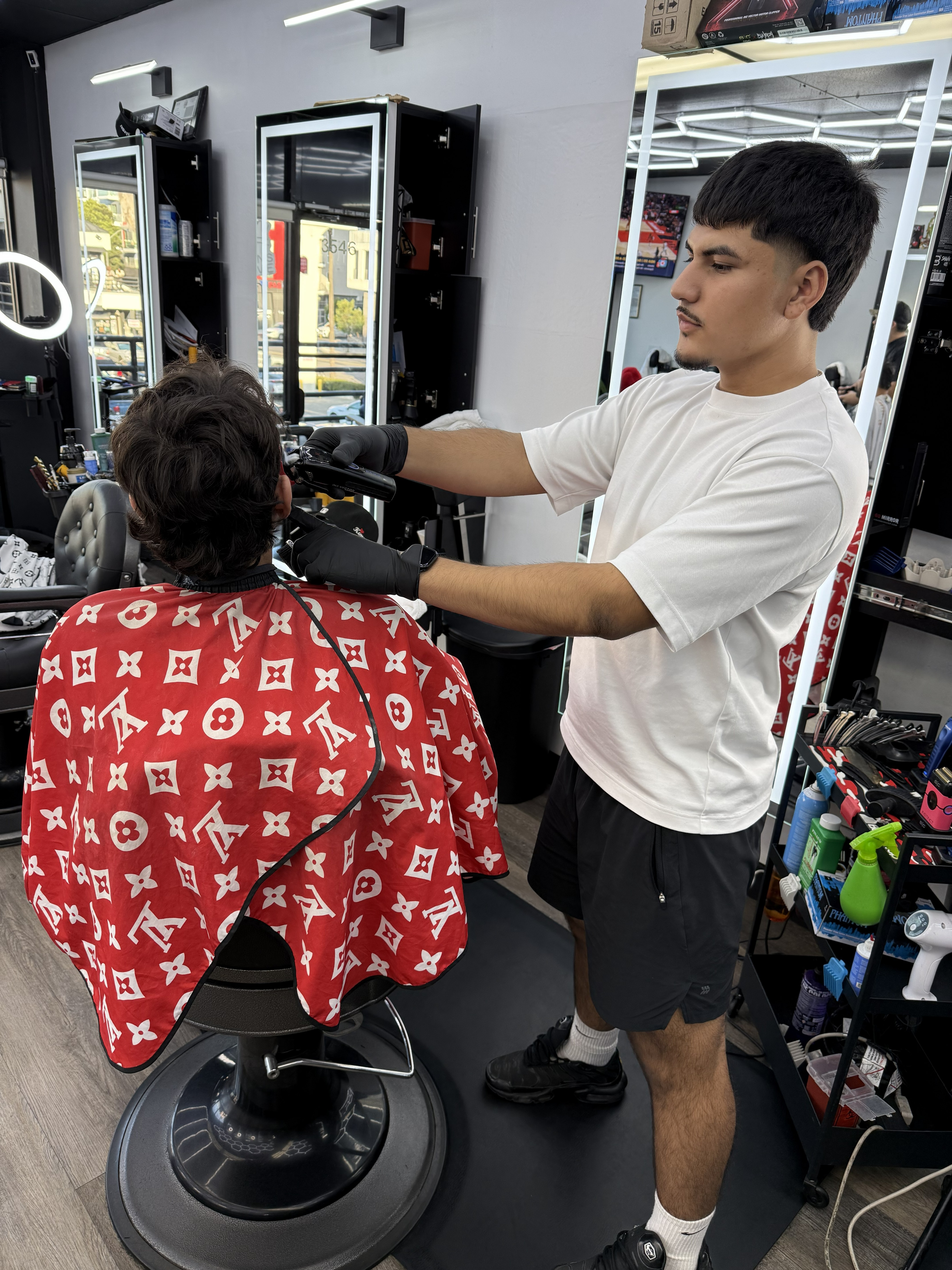 Barber standing with arms crossed in a modern barbershop, wearing a black cap, black T-shirt, and white pants, with hexagon ceiling lights in the background.