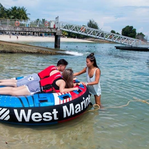 三个人在岸边；两个人躺在“West Marine”充气管上，第三个人在浅水中稳定它。