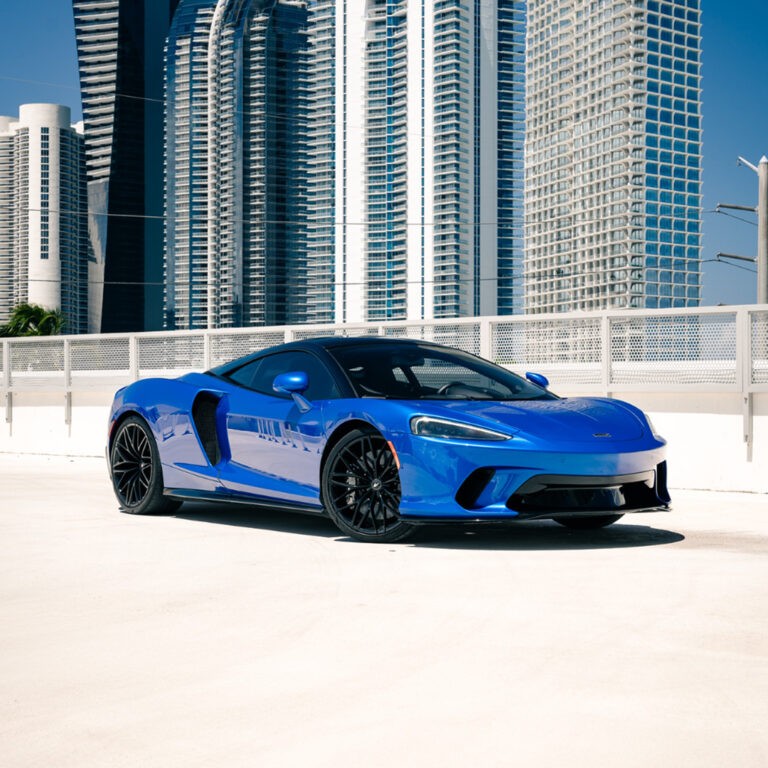 Front 3/4 view of a stunning blue McLaren GT for rent in Miami, parked with a modern skyline.