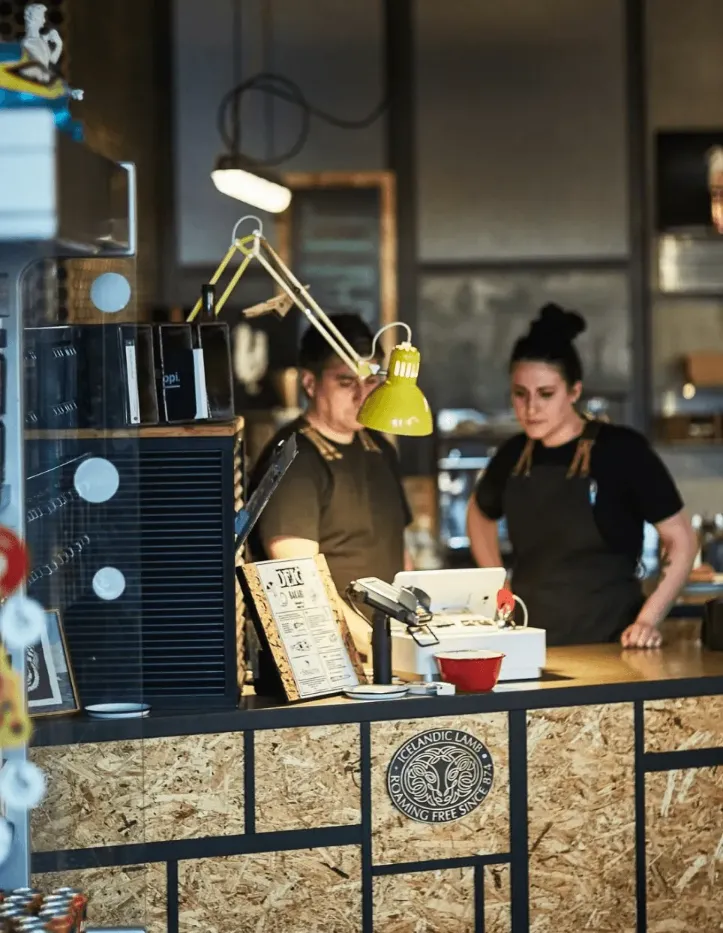 Two DEIG bakery staff members standing behind a service counter with a coffee machine, menu, and Icelandic lamb sign.